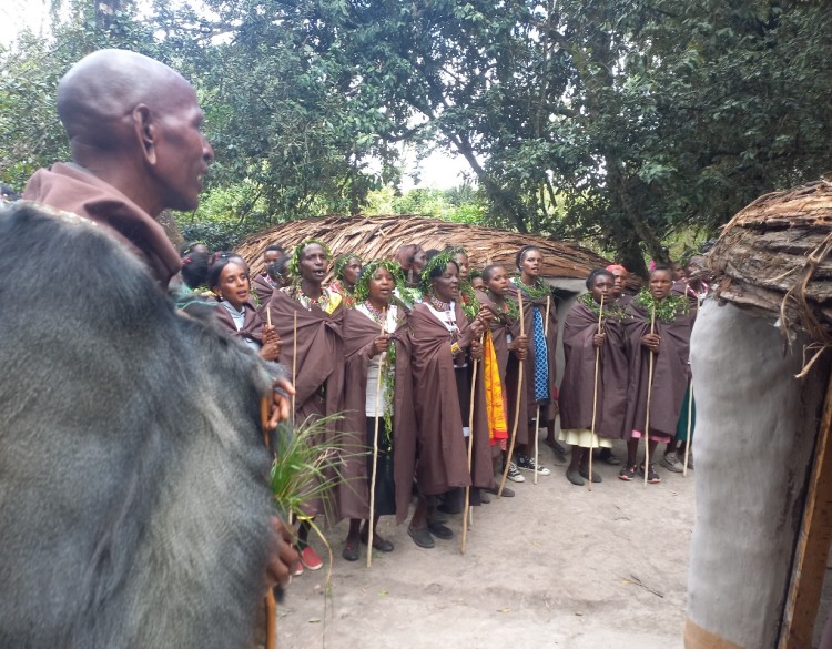 A group of Ogiek people, most of whom are wearing crowns or necklaces made of green leaves, stand in a line in front of a thatched roof building. Their mouths are open as if talking or singing.