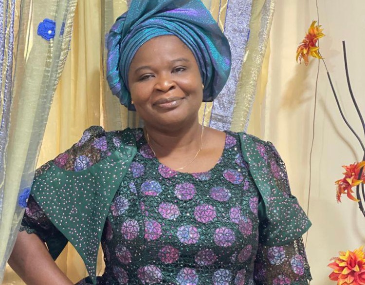 Dr. Bolanle Arokoyo, wearing a teal head wrap and a green and purple dress, smiles in front of a gold-colored curtain