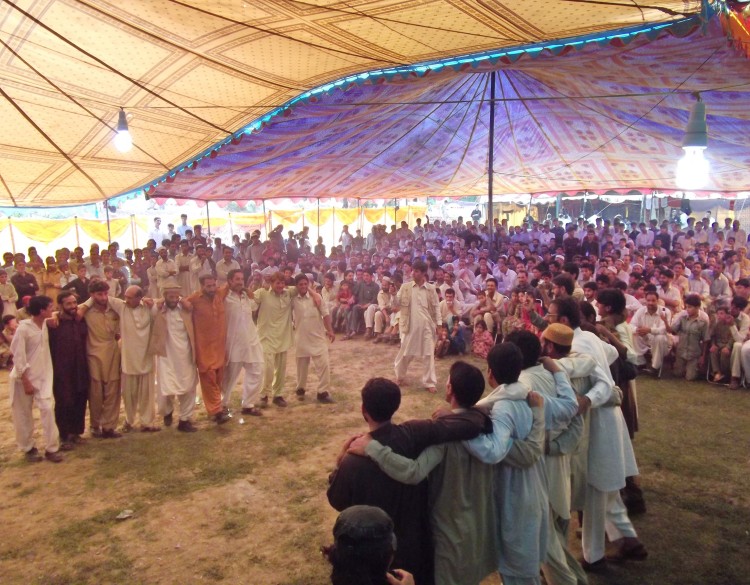 Two groups of men shoulder-to-shoulder face each other in traditional dance for a crowd under a large cloth tent.   Torwali dancers performing the traditional dance Dhiz (ɖʰiz) in the Simam Festival 2011. 