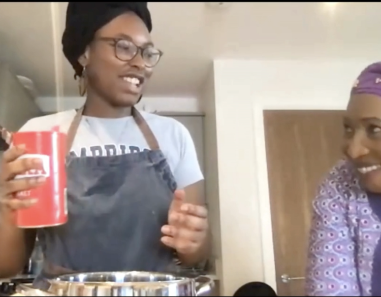 A young Nigerian woman holding a salt shaker above a large bowl, looking to the right at an older Nigerian woman, who is looking back at her and smiling.