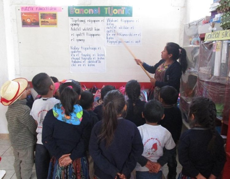 A group of 5-year-old Kaqchikel students look up at words written on a board, as their teacher stands at the front of the group and points to words on the board.