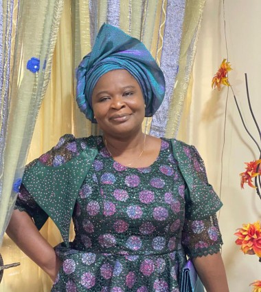 Dr. Bolanle Arokoyo, wearing a teal head wrap and a green and purple dress, smiles in front of a gold-colored curtain
