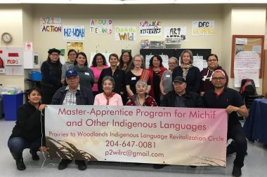 Photo of a group of 17 people in a classroom, holding up a banner which reads "Master-Apprentice Program for Michif and Other Indigenous Languages, Prairies to Woodlands Indigenous Language Revitalization Circle."