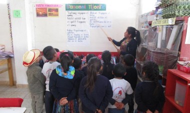 A group of 5-year-old Kaqchikel students look up at words written on a board, as their teacher stands at the front of the group and points to words on the board.