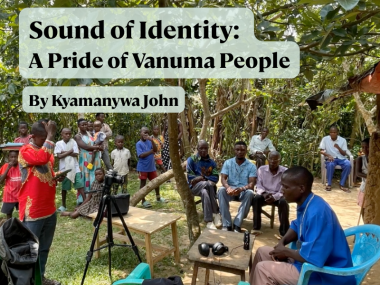 A group of twenty Vanuma people gathered under a large mango tree. To the left, a young man is looking down at an audio recorder, and to the right, an older man is speaking into a microphone. Between them, a video camera on a tripod is pointed at the older man.