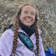 A young woman with two long braids, standing on large rocks on a beach and smiling