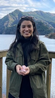 A Ryukyuan woman in a green coat, standing in front of a calm lake and green mountains, smiling