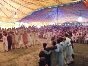Two groups of men shoulder-to-shoulder face each other in traditional dance for a crowd under a large cloth tent.   Torwali dancers performing the traditional dance Dhiz (ɖʰiz) in the Simam Festival 2011. 
