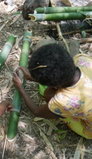 An Ayta Magbukun woman in a yellow shirt with short curly hair sits on the ground cutting bamboo stalks. 