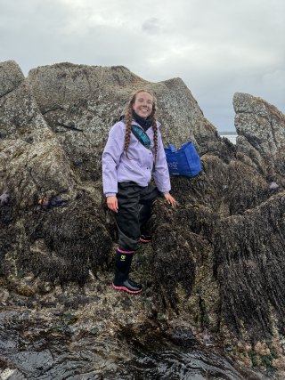 A young woman with two long braids, standing on large rocks on a beach and smiling