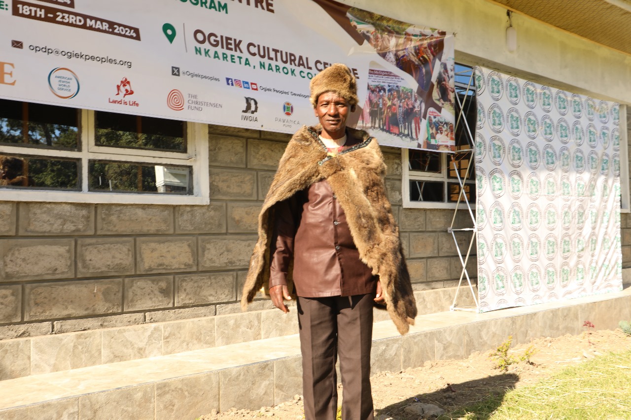 An Ogiek man wearing a fur cap and cape stands in front of the Ogiek Cultural Center. He is smiling faintly.