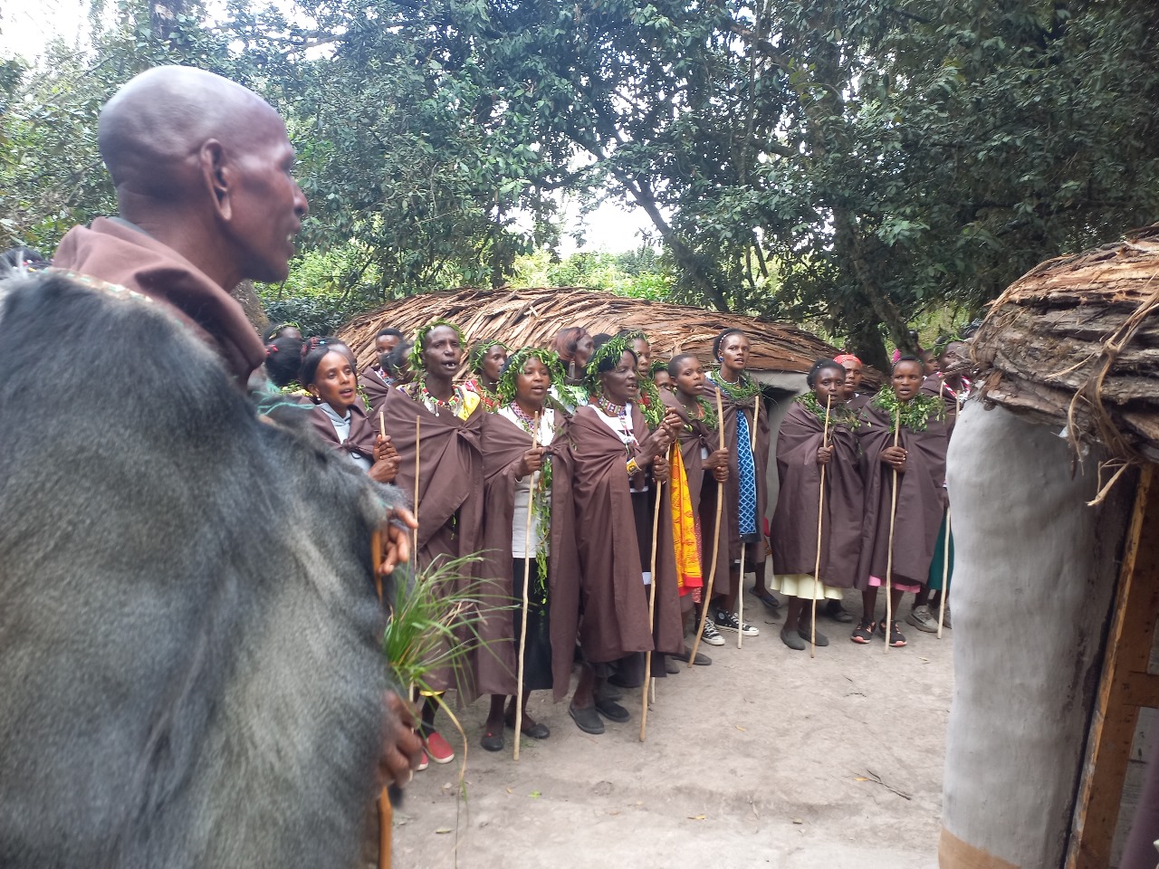 A group of Ogiek people, most of whom are wearing crowns or necklaces of green leaves, stand in a line in front of a thatched-roof building. Their mouths are open as if talking or singing.