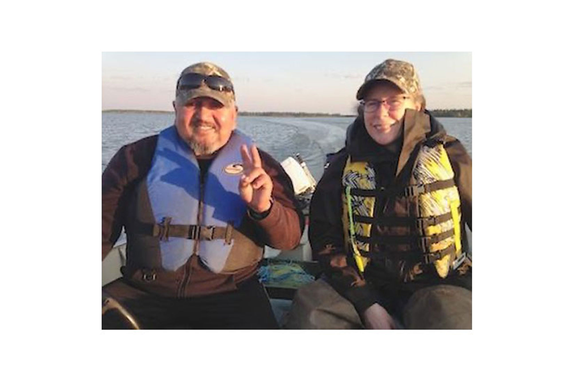 A photo of a man and a woman on a boat. They are wearing life jackets and camouflage hats, and smiling at the camera. The man is making a peace sign.
