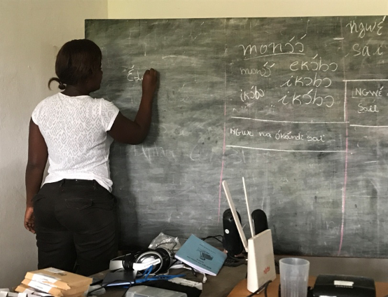 A young woman in a white shirt, writing in Iyasa on a chalk board, her back turned to the camera