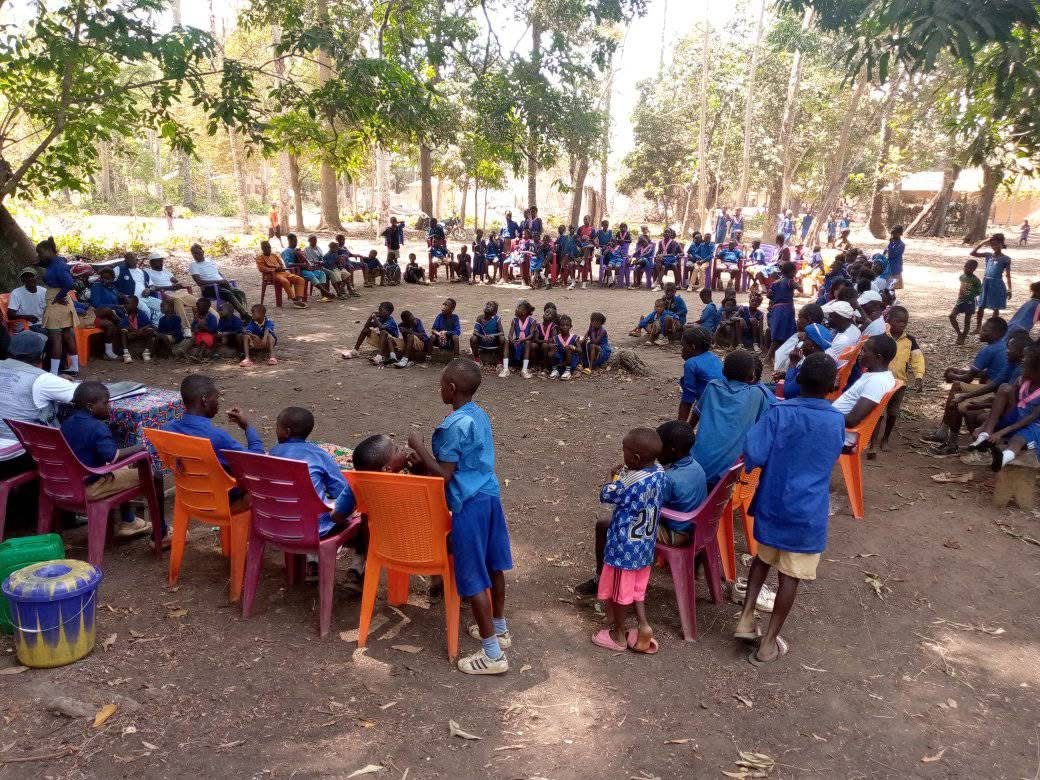 A group of school children and adults gathered under a large tree, many sitting on orange plastic chairs