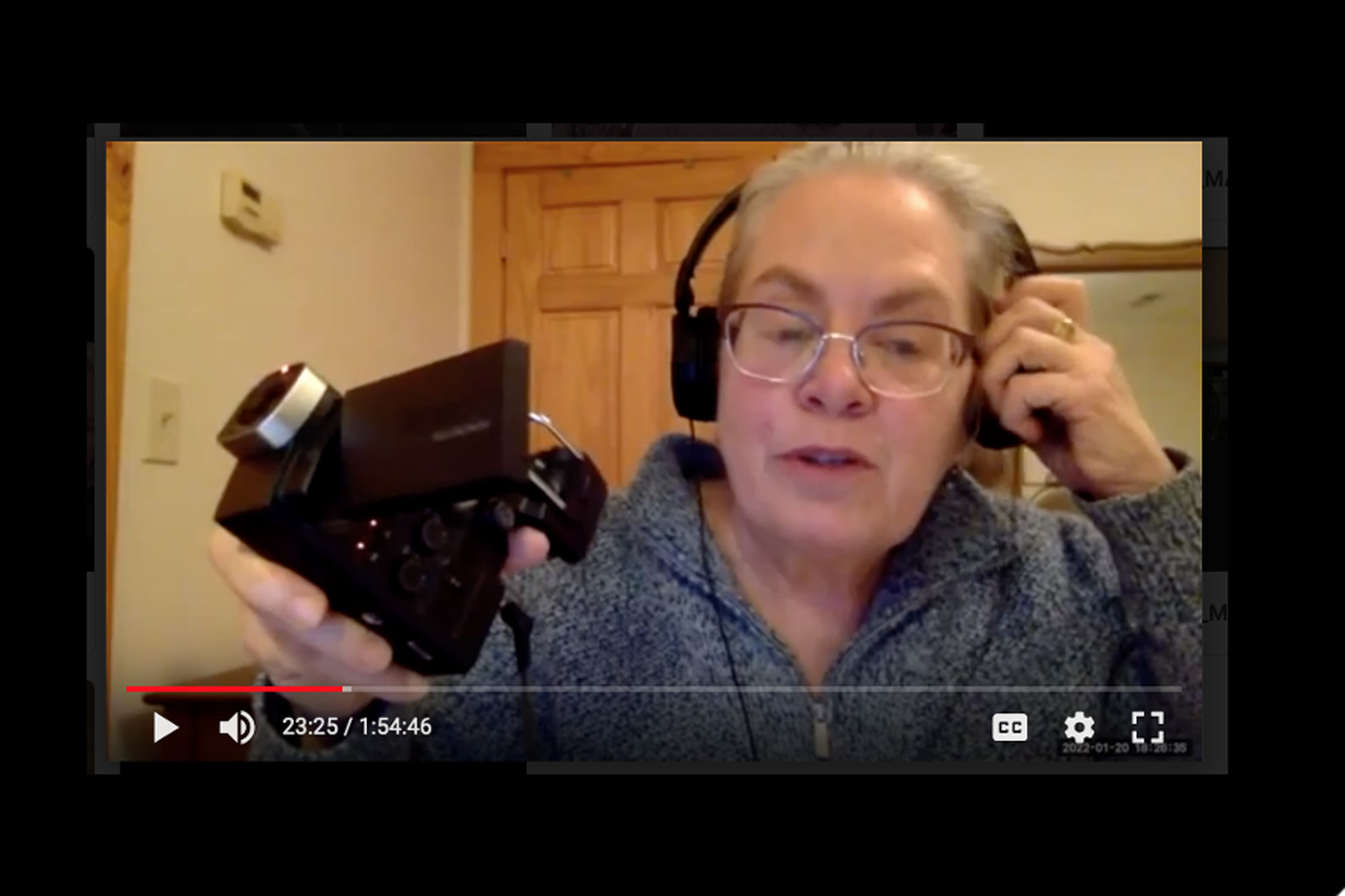 A video screenshot of a woman with grey hair, wearing headphones and glasses, and holding a video camera up to the viewer.