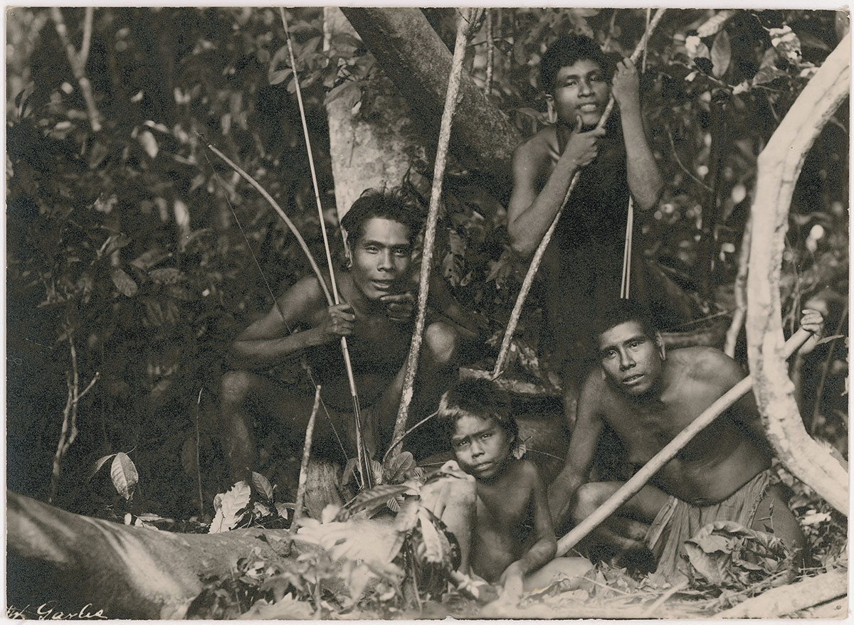 Black and white photo of three Krenak men and a small child kneeling in a forest. The three men are holding bows and arrows. 