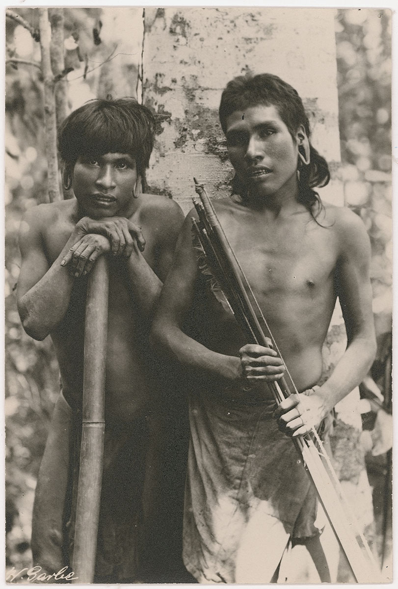 Black and white photo of two Krenak men leaning against a tree and holding bows and arrows.