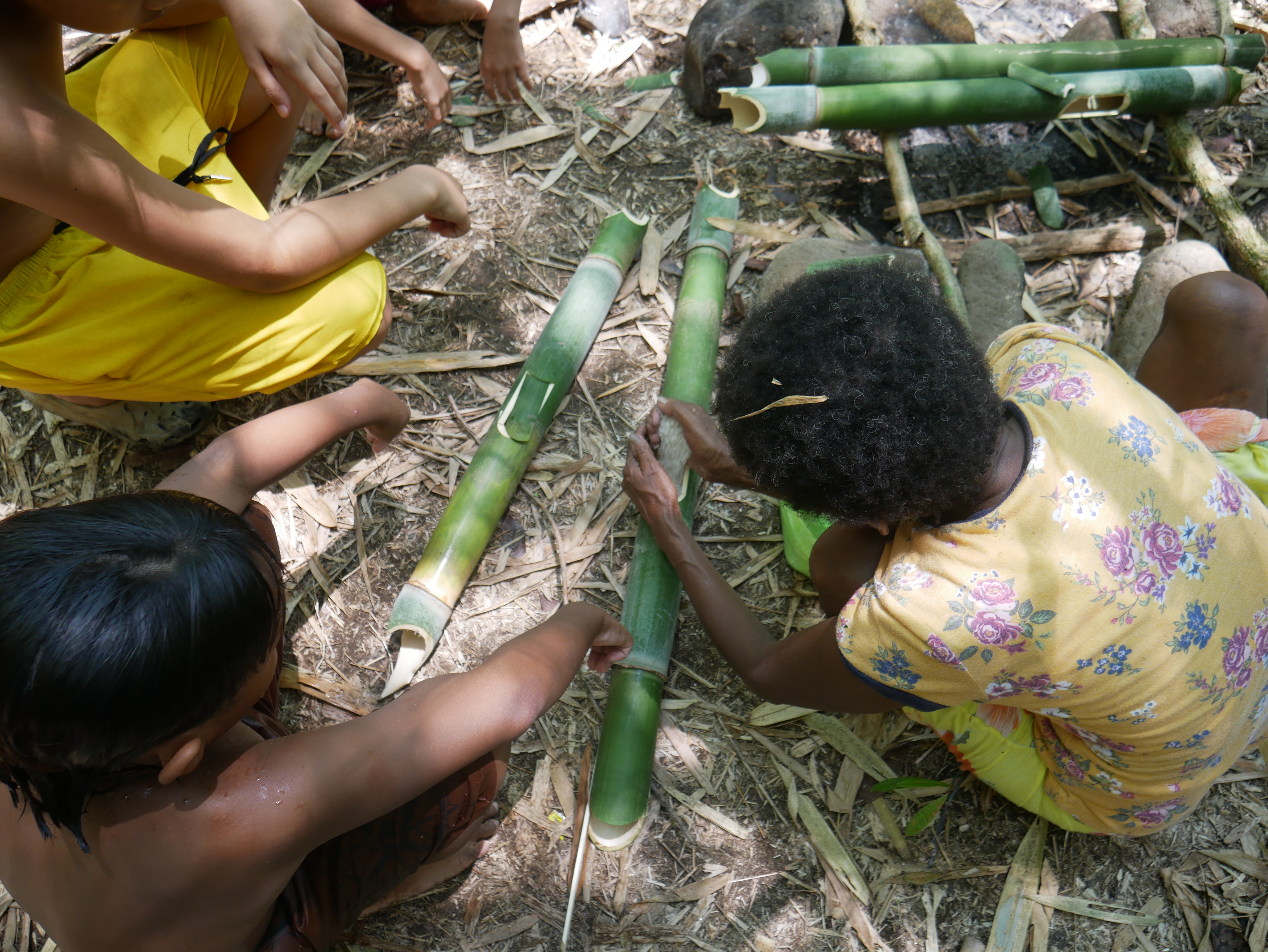 A group of people kneeling on the ground in the forest, watching a woman shape a bamboo pole.