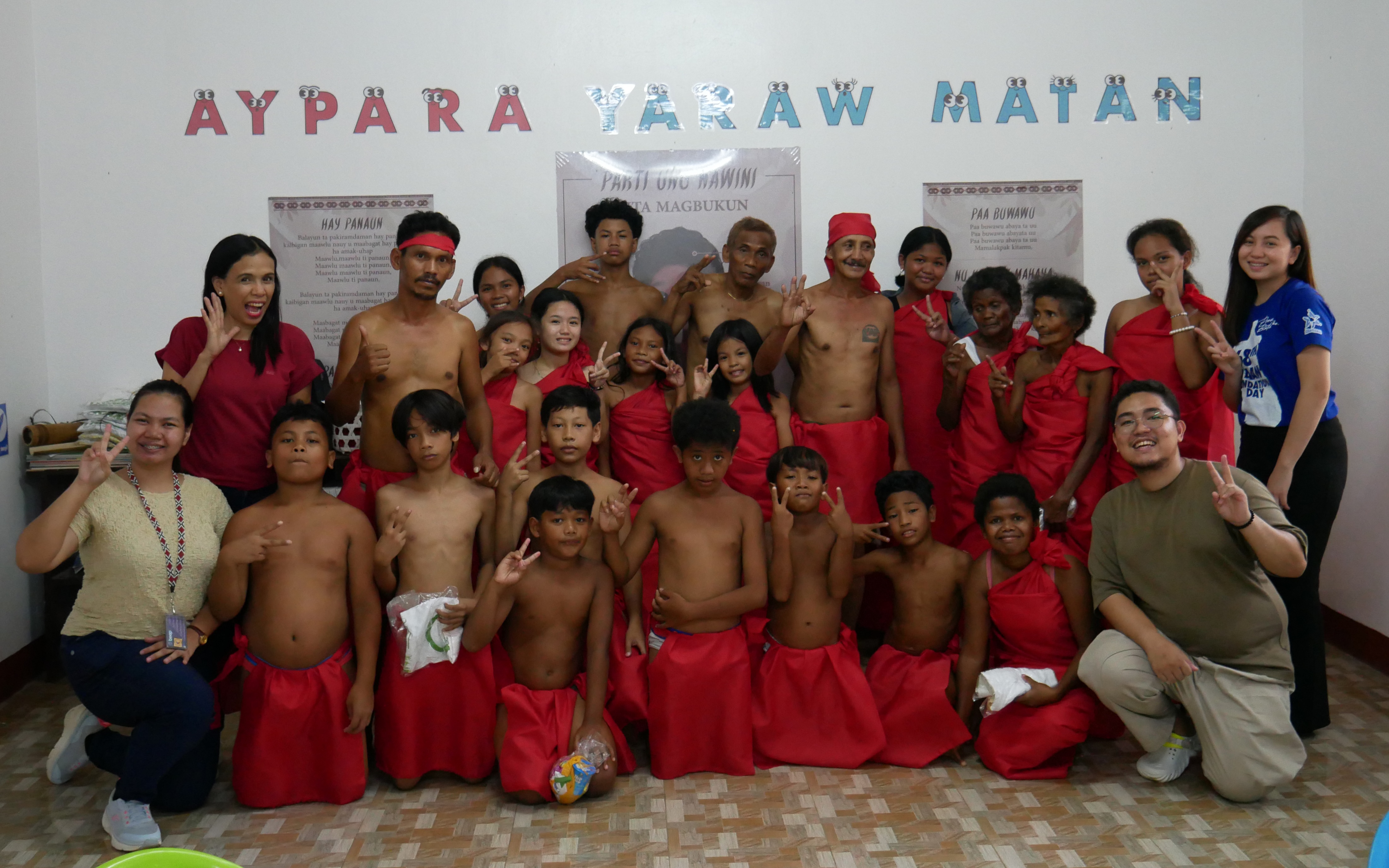 A group of adults and children posing in front of a wall with the words Aypara Yaraw Matan, along with posters showing vocabulary in the Ayta Magbukun language. Most of the people are wearing red tradtitional Ayta garments.