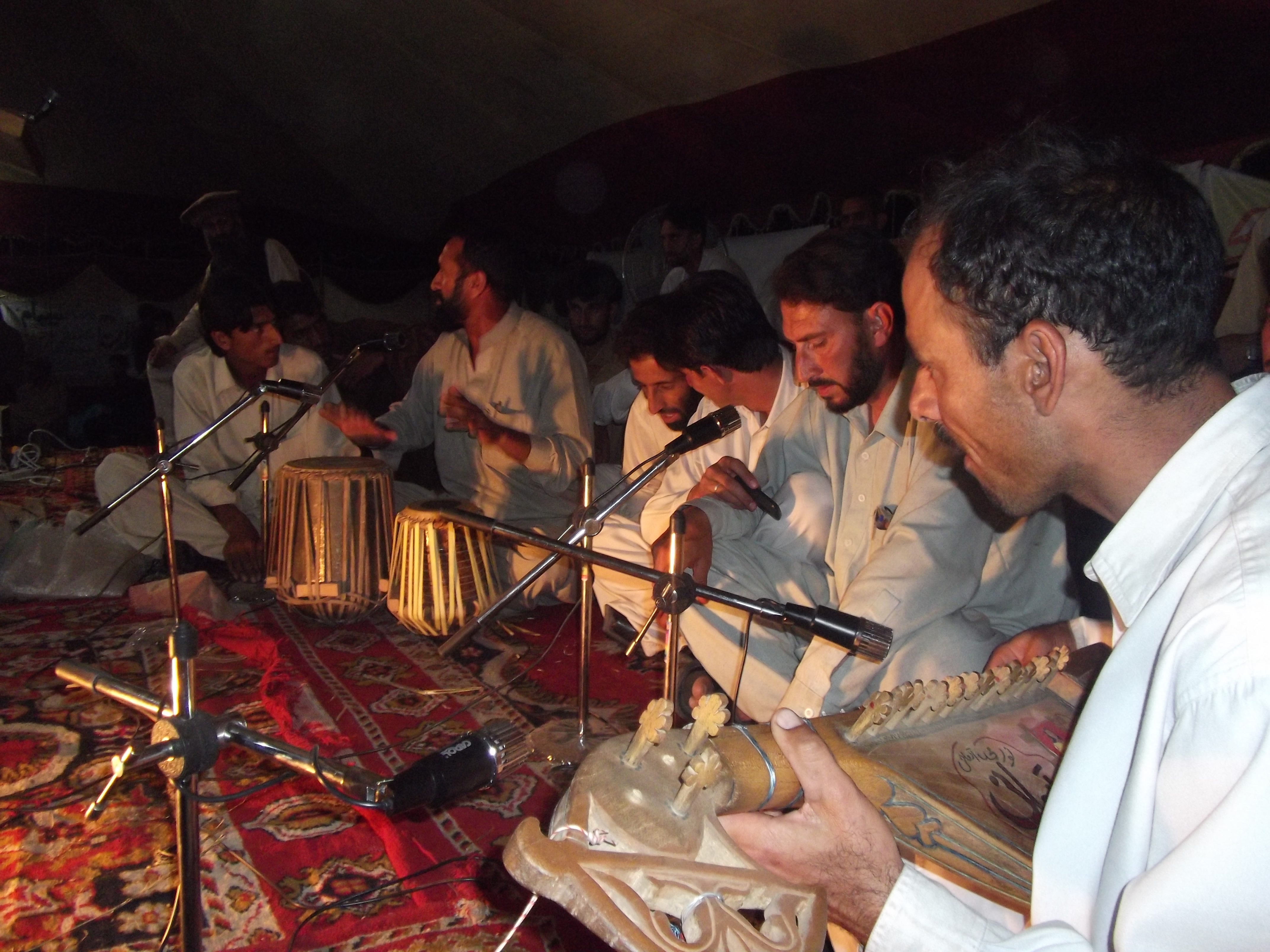 A group of torwali men, seated on the ground on an ornate red carpet, play drums and stringed instruments and sing into microphones in front of them.