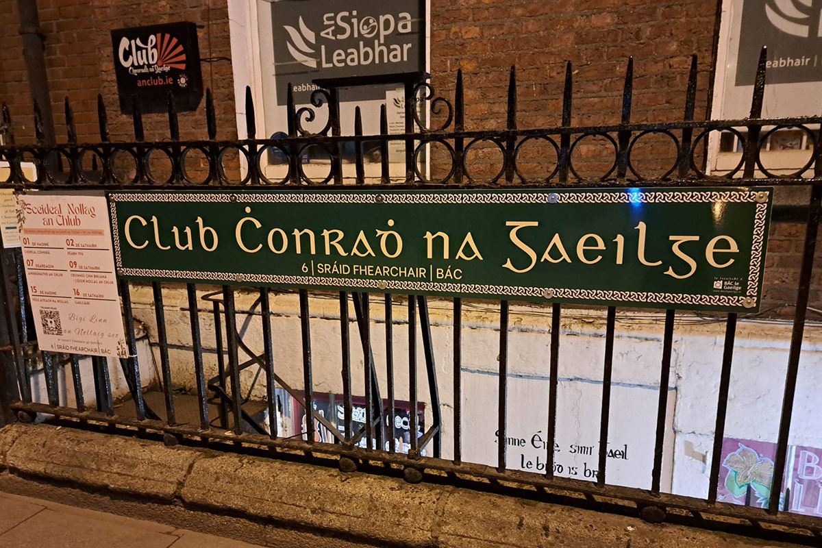 A dark green sign with gold lettering reading "Club Chonradh na Gaeilge". The sign is hung on an iron fence outside a brick building.