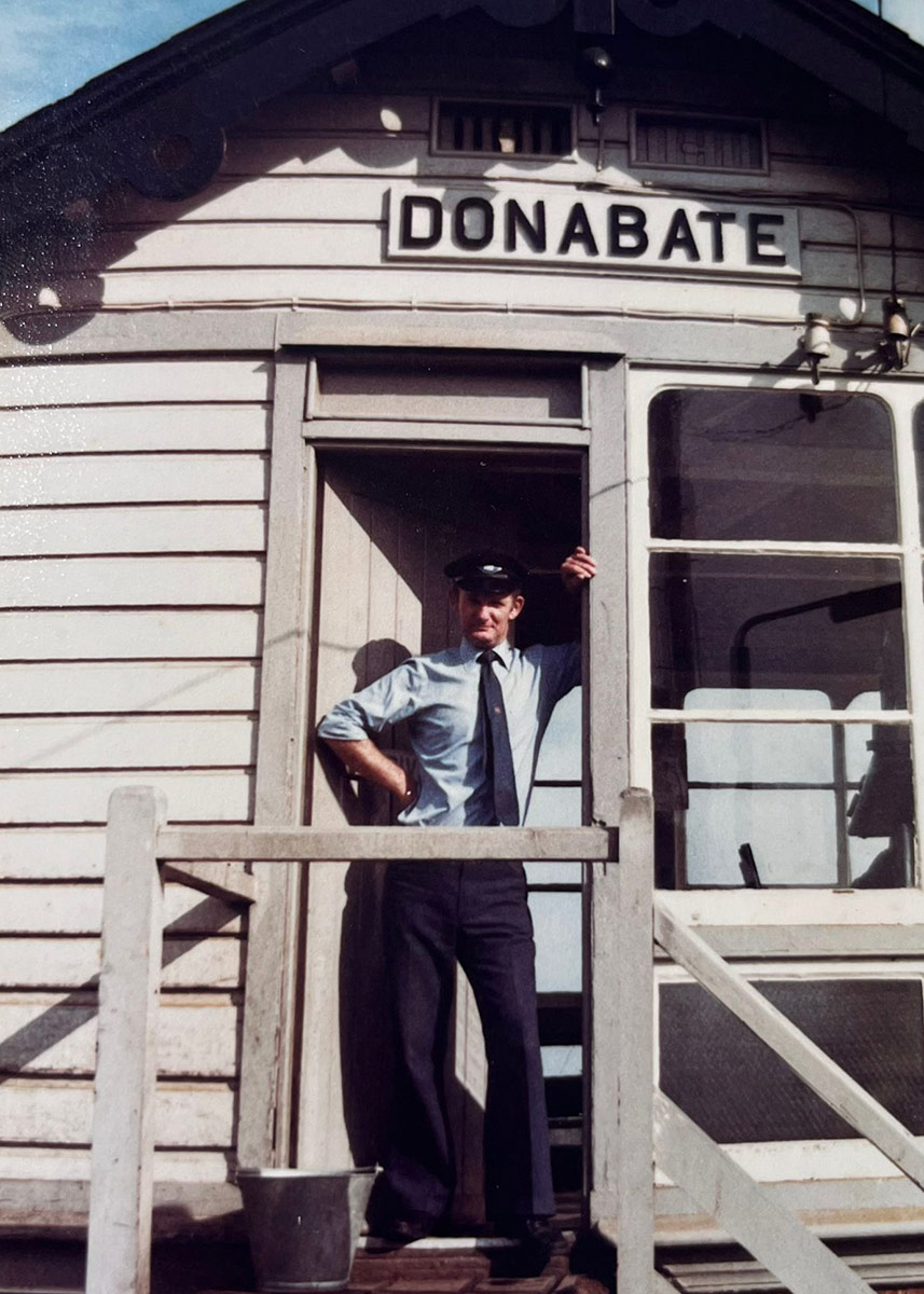A man in a blue railway uniform under a sign reading "DONABATE". Old color photograph.