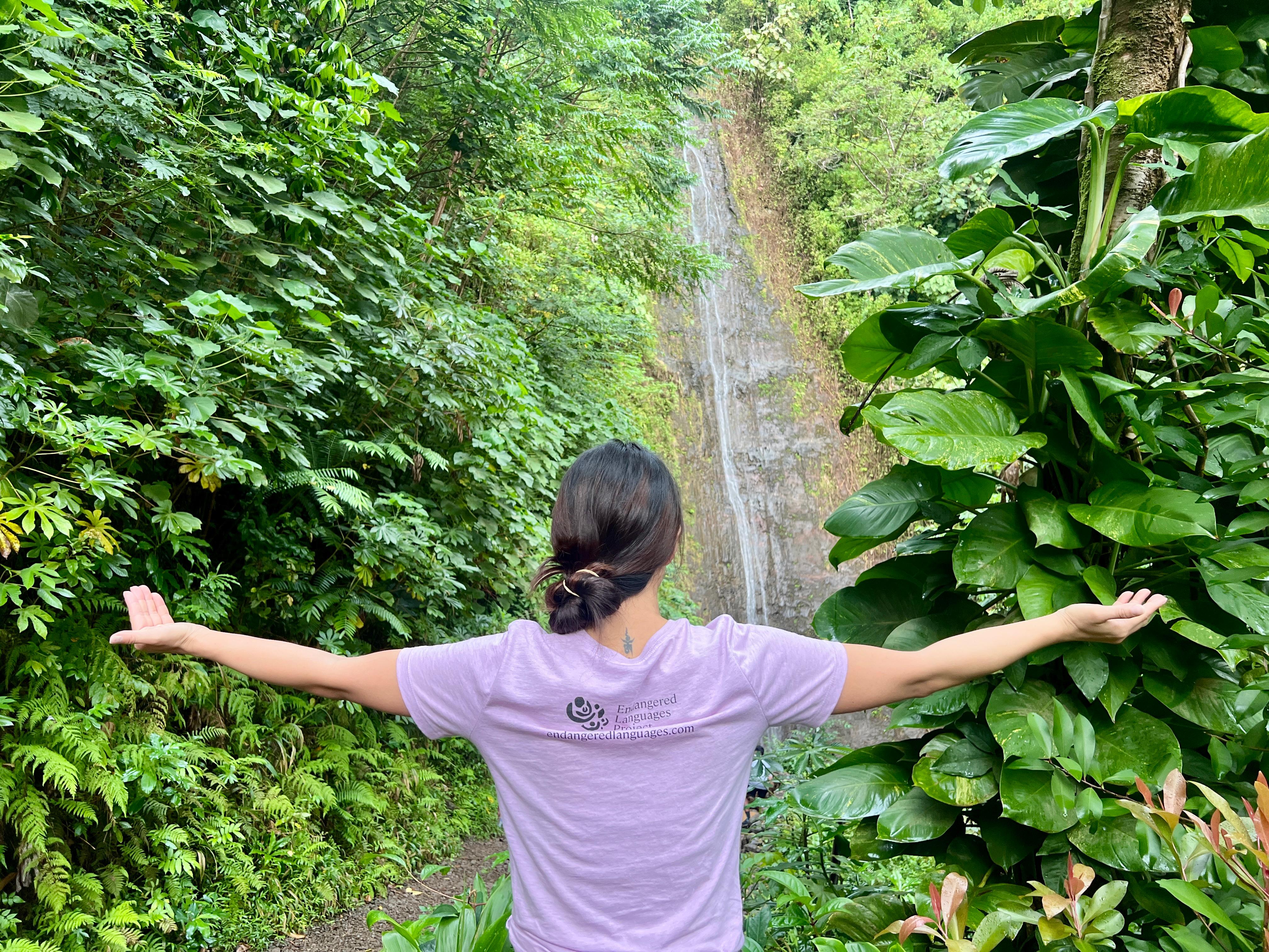 A woman in a pink ELP shirt raises her arms, palms up, in front of a waterfall surrounded by green plants.