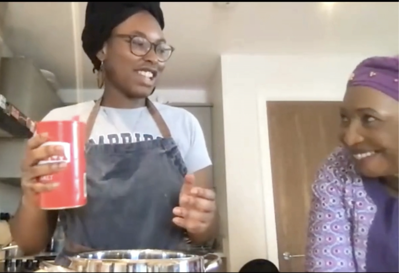 Beulah Waritimi, a young Nigerian woman, holds a salt shaker over a bowl. She is looking to the right at an older Nigerian woman, her grandmother, who is smiling back at her.