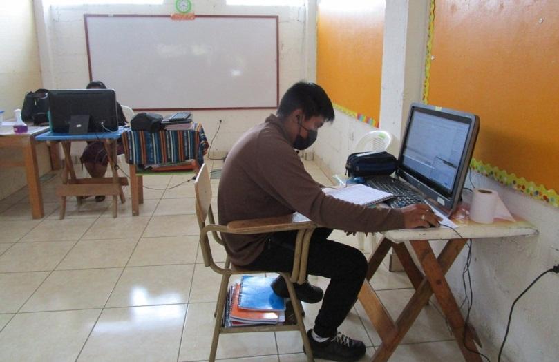A student sitting at a computer in a classroom. He is wearing a mask and looking down at a notebook.