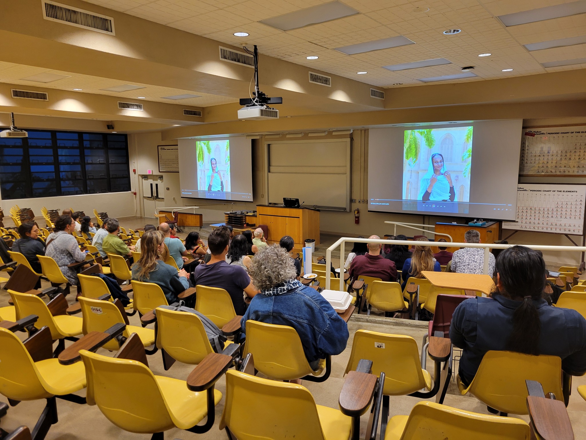 A group of people in an auditorium look at a projector screen showing an image of a young Sudanese woman in a white headscarf.