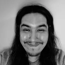 A black and white photo of a young Cree man with long dark hair and a mustache and goatee, smiling at the camera