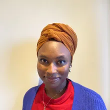 A young Nigerian woman in a brown head wrap, red shirt, and blue cardigan, smiling at the camera