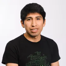 A young Quechua man with short hair, wearing a black t-shirt, and smiling at the camera in front of a grey background.