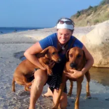A woman kneeling on a beach hugging two brown dogs.