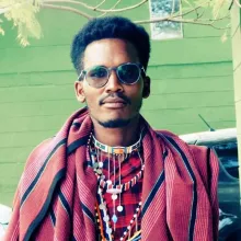 A young Maasai man with a mustache and goatee. He is wearing sunglasses, beaded necklaces, and a red wool shawl, standing in front of a green wall.