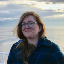 A woman with long reddish hair and large glasses, smiling in front of a sunset over the Salish Sea.