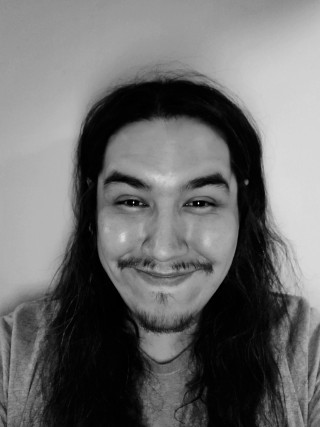 A black and white photo of a young Cree man with long dark hair and a mustache and goatee, smiling at the camera