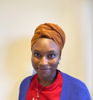 A young Nigerian woman in a brown head wrap, red shirt, and blue cardigan, smiling at the camera