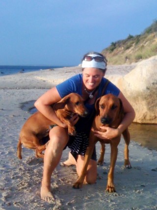 A woman kneeling on a beach hugging two brown dogs.
