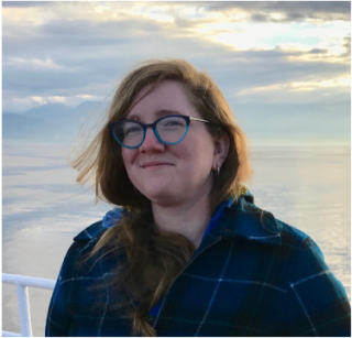 A woman with long reddish hair and large glasses, smiling in front of a sunset over the Salish Sea.
